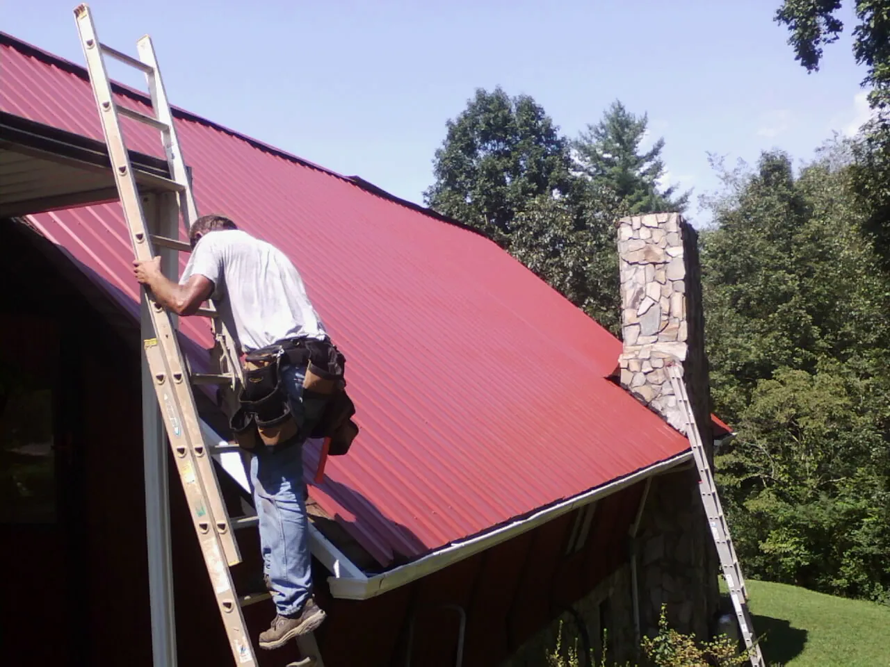 Roofer on a ladder beside a red metal roof and stone chimney — Soffit & Fascia Repair by Roofing Brothers in El Dorado